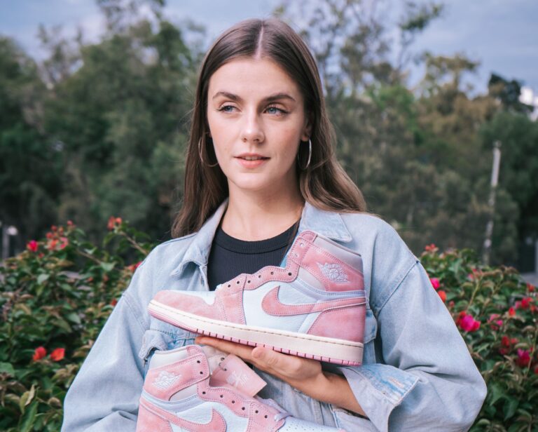 Stylish woman in a park holding pink Nike sneakers, wearing denim outfit.